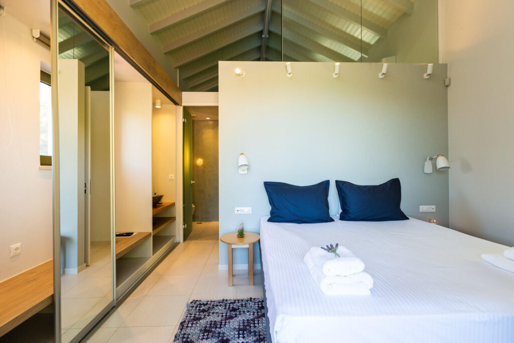 A modern minimalist bedroom in a luxury vacation home, featuring a king-size bed with crisp white linens, two navy blue pillows, and neatly folded white towels. Sliding mirrored wardrobe doors open to a walk-in closet area with a wooden shelf. A small wooden side table holds a potted plant. The room has high exposed wooden beam ceilings, soft recessed lighting, wall-mounted lamps, light green walls, tiled floors, and a colorful woven rug.