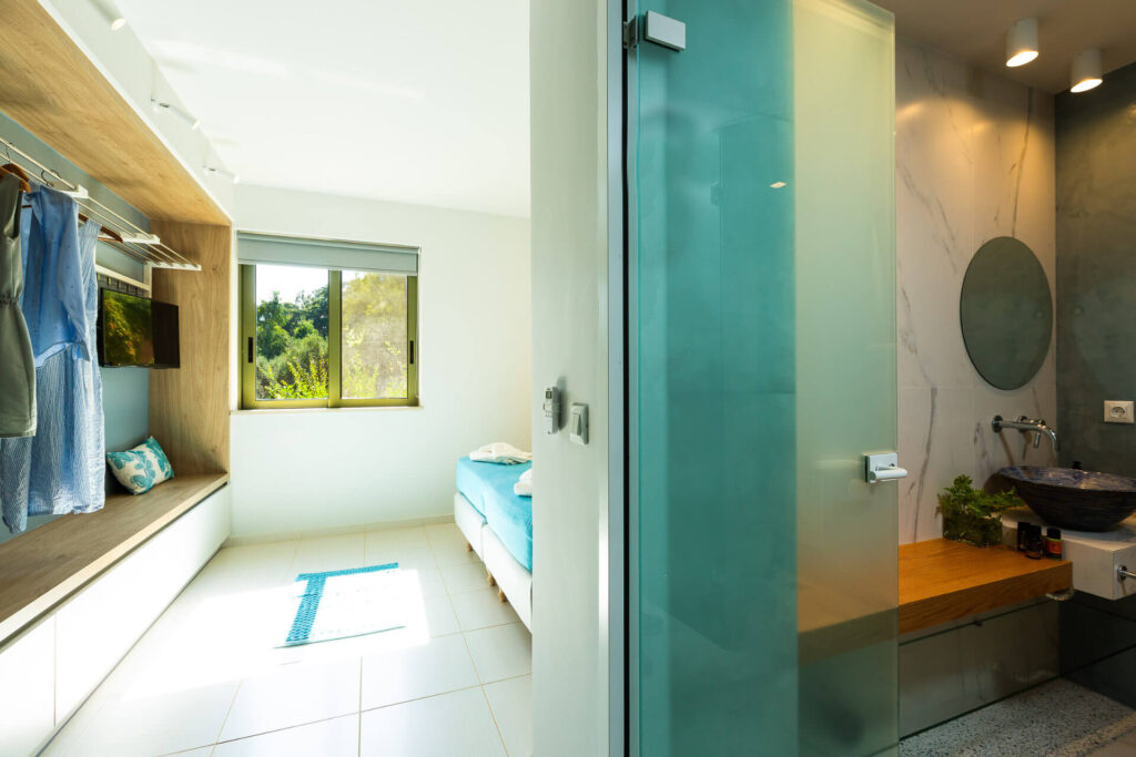 A modern minimalist guest bedroom in a luxury vacation home, featuring a bed with a blue mattress and white linens. Light blue clothing hangs on wooden hangers in a built-in open wardrobe with shelves. A cushioned window bench in blue overlooks lush green trees through a large window with sheer blinds. A frosted glass partition door opens to a stylish bathroom with a round mirror, white sink, wooden accents, and plants. The room has white walls, tiled floors with sunlight streaming in, recessed lighting, and contemporary design elements.