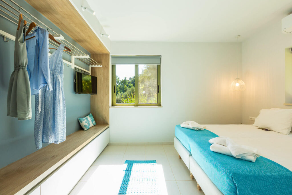 A modern minimalist guest bedroom in a luxury vacation home, featuring a bed with turquoise linens and neatly folded white towels. Light blue clothing hangs on wooden hangers in a built-in open wardrobe with shelves. A cushioned window bench in blue overlooks lush green trees through a large window with sheer blinds. The room has white walls, tiled floors with sunlight streaming in, recessed lighting, a wall-mounted lamp, and contemporary design elements.