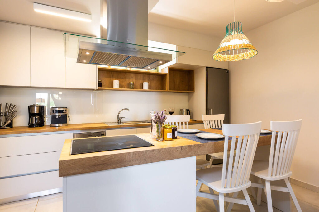 A modern minimalist kitchen in a luxury vacation home, featuring a spacious white island countertop with a wooden top, built-in induction cooktop, and bar seating with white wooden stools. Glossy white cabinets, wooden shelves, a glass-fronted stainless steel range hood, integrated refrigerator, sink, coffee maker, and utensils are visible. A pendant light hangs above, with fresh lavender and oil bottles on the counter, set against tiled floors and neutral walls.
