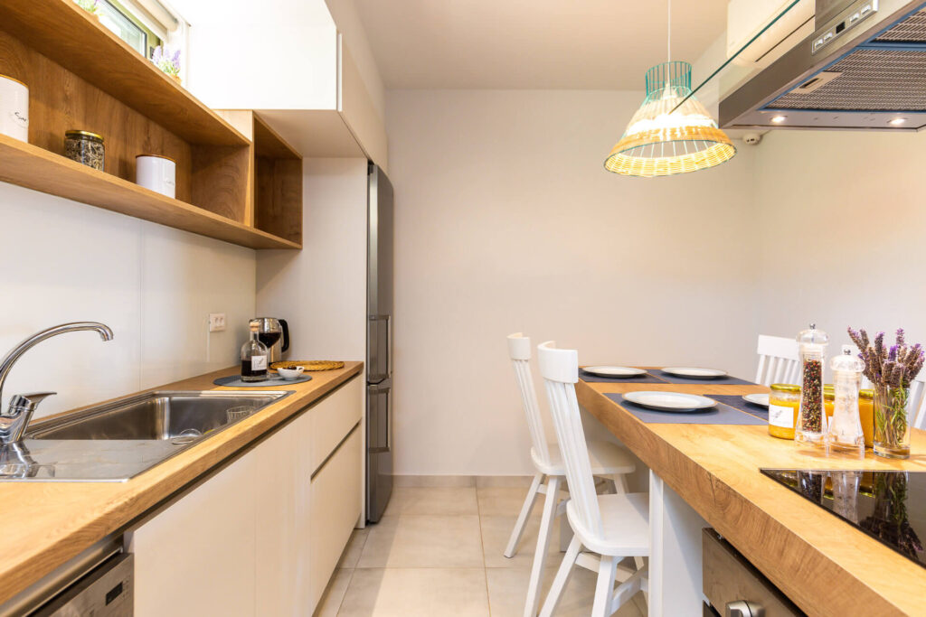 A modern minimalist kitchen in a luxury vacation home, featuring a wooden-topped countertop with sink, induction cooktop, and bar seating with white wooden stools. Glossy beige cabinets, wooden open shelves with jars and cups, stainless steel range hood, integrated refrigerator, faucet, oil bottles, salt and pepper shakers, and fresh lavender are visible. A colorful pendant light hangs above, set against white walls and tiled floors.