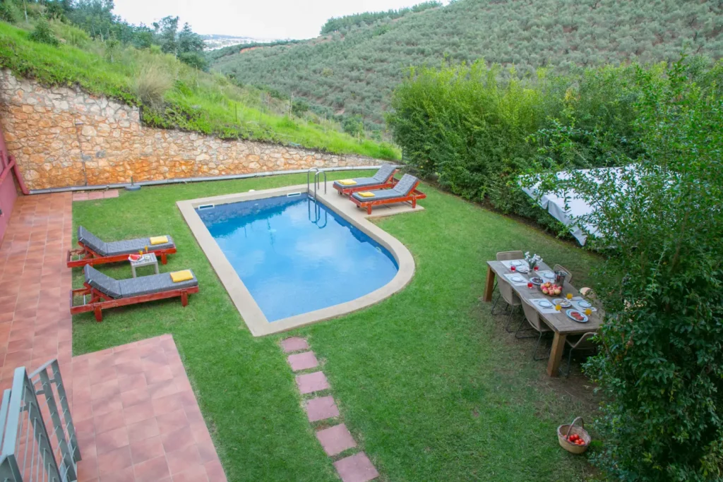 A private garden with a rectangular swimming pool, four sun loungers with yellow towels, and a wooden outdoor dining table set for a meal, surrounded by green hills and stone walls.