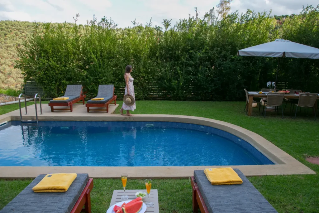 A woman in a white summer dress holding a hat walks beside a blue swimming pool with sun loungers, drinks, and a nearby outdoor dining table under a large umbrella, surrounded by greenery.